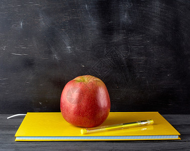 红色成熟的苹果躺在黄色封闭笔记本上黑色粉笔板的背景图片