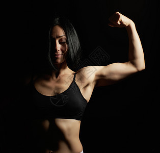 美丽的年轻运动女孩举起并弯下她的手臂展示Biciceps运动员站在黑暗背景上穿着黑色的衣和短裤图片