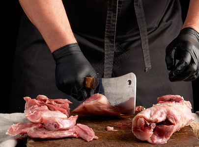 用黑色乳胶手套的厨师拿着一把大刀在棕色木制板上切碎生兔肉在黑暗背景下做饭图片