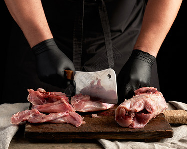 用黑色乳胶手套的厨师拿着一把大刀在棕色木制板上切碎生兔肉在黑暗背景下做饭图片