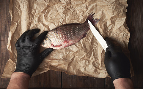 戴着黑手套的厨师在一张棕色的纸上清洗整条鲤鱼的鳞片俯视图图片