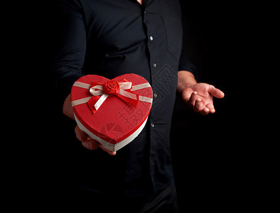 穿黑衬衫的成人男子持有红纸板盒其形式为心脏在黑暗背景上鞠躬有节日问候惊喜和礼物的概念图片