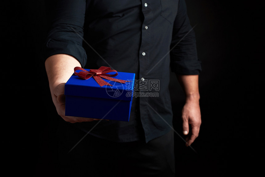 穿黑衬衫的成年男子拿着一个蓝色的方形盒子红弓绑在黑暗背景上假日问候的概念惊喜图片