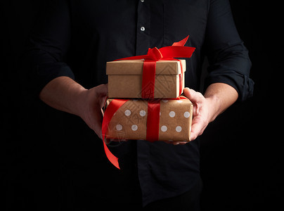 穿黑衬衫的成年男子拿着一堆包棕纸的礼物黑色背景的红弓假日问候惊喜和礼物的概念图片