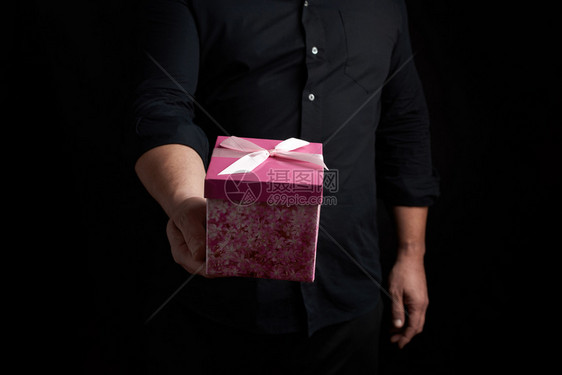 穿黑衬衫的成年男子拿着粉红色的方形盒子弓绑在黑暗背景上祝贺的概念惊喜图片