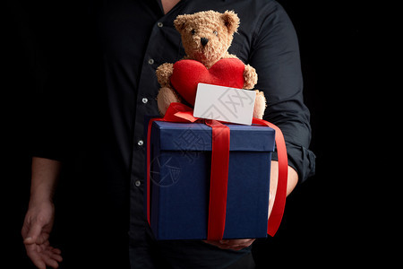 身穿黑衬衫的成人男子拿着一个蓝色广场盒子绑着红色丝带坐在棕泰迪熊顶上图片