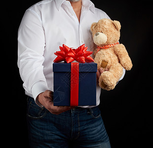 2月14日一名身穿白衬衫的成年男子手持一个蓝色方形盒子盒子上系着红丝带和棕色泰迪熊表达对情人节的祝贺图片