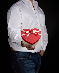 穿白衬衫的成人男子持有红纸板盒其形式为心脏在黑暗背景上鞠躬有假日惊喜和礼物的概念图片