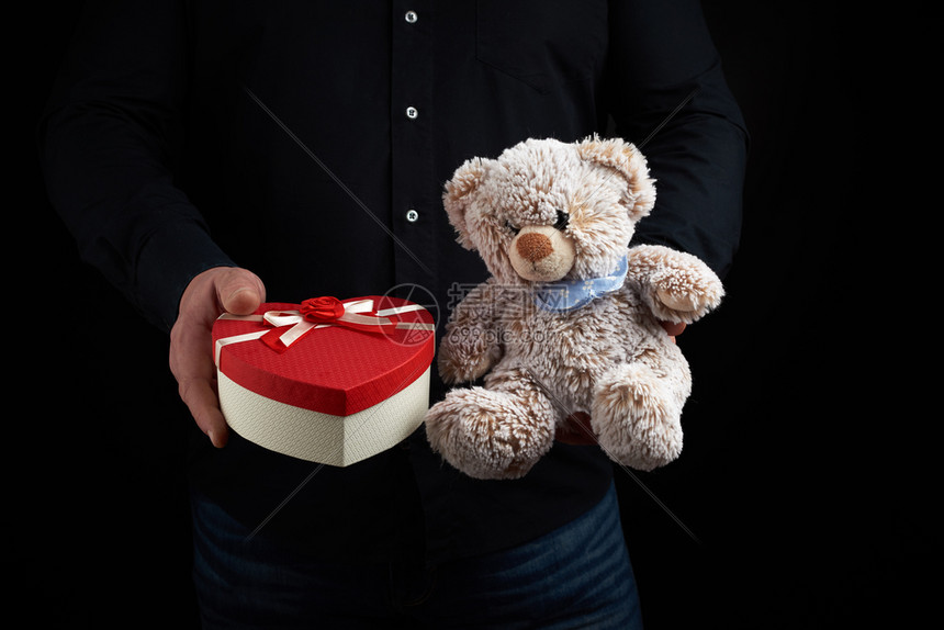 身穿黑衬衫的成人男子拿着一个蓝色方格盒子上面绑着红丝带和棕褐色泰迪熊图片
