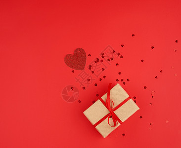以棕色克拉夫纸包裹的长方形盒子绑有红色薄丝带装饰闪亮的心在附近散落节日是情人红色背景图片