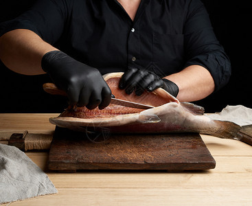 黑色制服和乳胶手套的厨师将一具新鲜鲑鱼切成黑色背景的木制剪板上图片