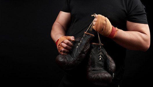 身穿黑色制服的肌肉运动员手上握着非常古老的棕色拳击手套双被绑上橙色弹运动绷带黑色背景复制空间图片