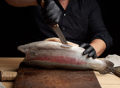 黑衬衫主厨和乳胶手套把一整条新鲜的鲑鱼切下来在旧棕色切面板上低键图片