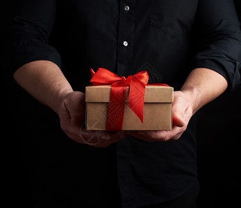 身穿黑衬衫的成人男子手上握着一个平方棕色的盒子上面绑着黑色背景的红丝带是节日问候惊喜和感激的概念图片