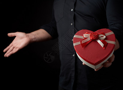 穿黑衬衫的成人男子持有红色纸板盒其形式为心脏在黑暗背景上鞠躬有假日惊喜和礼物的概念图片