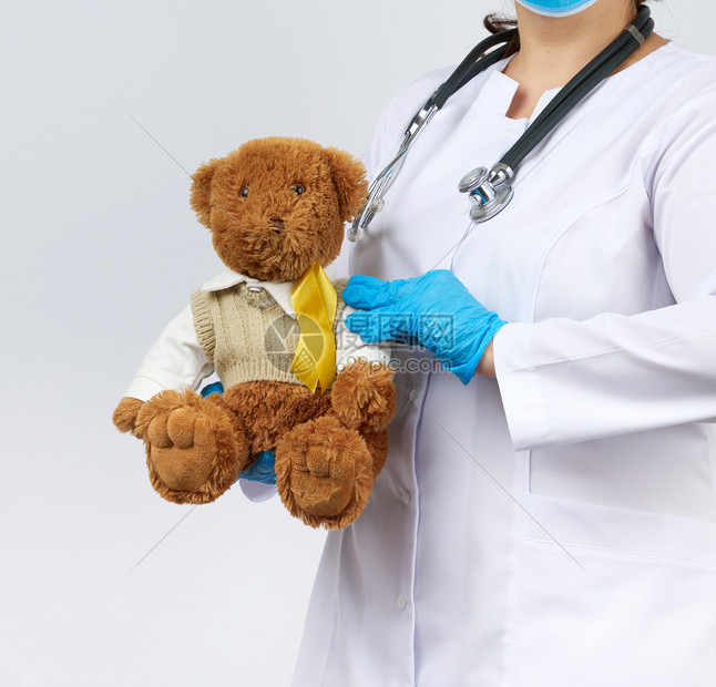 穿白外套的儿科医生,蓝乳胶手套戴着棕色泰迪熊,毛衣上有黄丝带,与儿童癌症作斗争的概念,问题图片