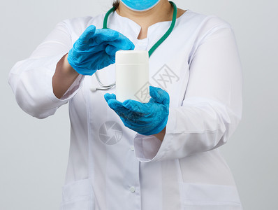 身穿白色大衣和面罩的女医生戴白色塑料罐装药品和手持片的白色塑料罐戴蓝乳胶手套白底图片