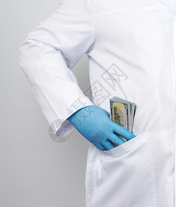 穿白色医疗大衣和蓝乳胶手套的男医生将一堆美元放在他的外套口袋里这是贿赂图片