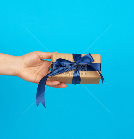 手握着一个盒子礼物包在棕色的Kraft纸上绑在蓝色背景的丝绸带上赠送礼物的概念图片