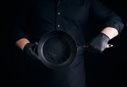 穿黑色制服和乳胶手套的男厨师在前面拿着一个空的圆旧黑色铁铸板低键背景图片