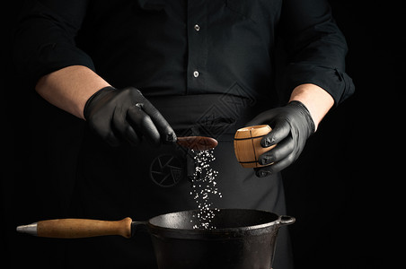 黑制服和乳胶手套的男烹饪用黑色铸铁锅制成盐类食品晶体在空气中冻低键图片