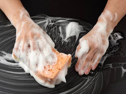 白色肥皂泡沫中的两个女手握着一湿海绵擦黑表面清洁和洗表面的概念顶视图片