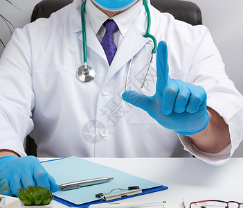 穿着白色大衣和蓝乳胶手套的医生坐在办公室的白工作桌前手举着注意的势用食指接受病人的概念图片