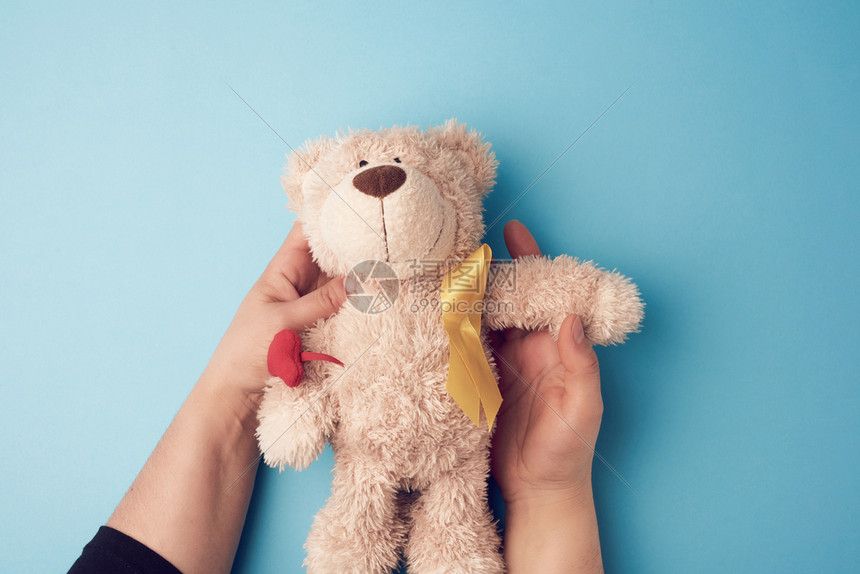 女性手握着一只小泰迪熊,黄丝带折叠在蓝色背景的循环中。 与儿童癌症作斗争的概念。 问题图片
