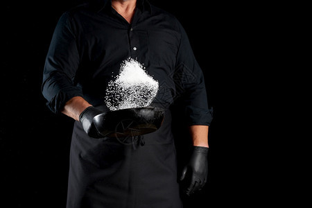 身穿黑色制服的男子拿着一个带盐的圆面铁锅厨师把白盐扔在黑色背景上图片
