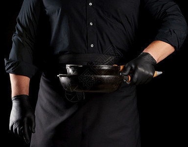 穿黑色制服和乳胶手套的男厨师在前面拿着一个空的圆旧黑色铁铸板低键背景图片