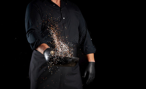 穿着黑色制服的男子拿着圆制铁锅配有盐和辣椒的铁锅黑背景上涂着辣椒的大厨复制空间图片