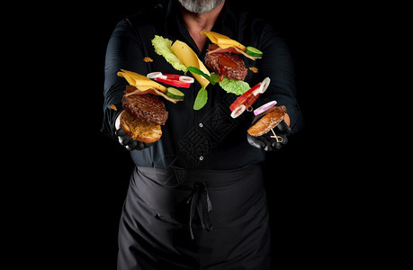 黑衬衫厨师围裙和乳胶黑手套站在色背景上手握着起司汉堡成份一包带芝麻种子切菜番茄生和洋葱环奶酪图片