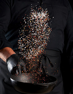 穿着黑色制服的男子拿着一个圆面铁锅配有盐和辣椒的铁锅黑背景厨师把香料扔起来图片