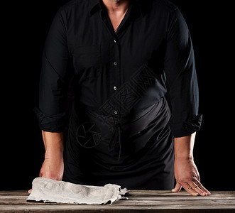 穿着黑衬衫和围裙的男厨站在木制桌旁手举起来黑色背景图片