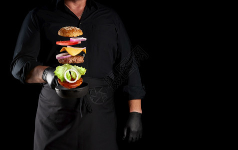 身穿黑色制服的成年男子持有铁环煎锅配冻奶酪汉堡成分芝麻面包奶酪番茄洋葱肉片黑色背景复制空间图片