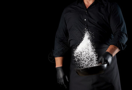 身穿黑色制服的男子拿着一个带盐的圆面铁锅厨师把白盐扔在黑色背景上图片