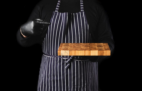身穿条纹蓝色围裙和黑衣服的男主厨站在黑色背景的对面手里握着一个长方形木制厨房切开板关上背景图片