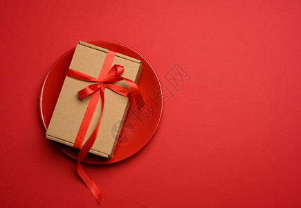长方形的棕色纸板箱上系着一条丝红色的丝带放在一个圆形的陶瓷红色盘子里俯视图图片