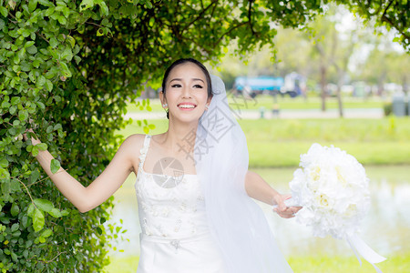 在婚礼当天美丽的年轻女在树墙上穿着白色礼服衣图片