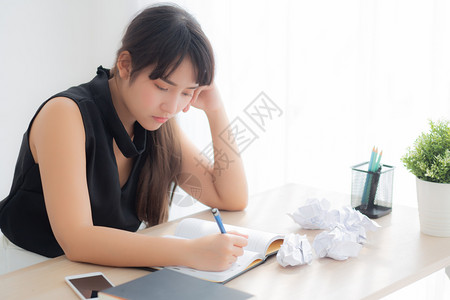 美丽的亚洲妇女疲惫不堪在办公桌上写作过量压力重女孩对办公室的笔记本和折叠纸自由职业者和商概念一无所顾虑图片
