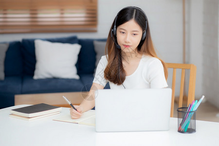 年轻女在网上戴耳机书桌上进行电子学习女孩戴耳机在家用笔记本电脑学习因特网接受新的正常教育远程和培训图片