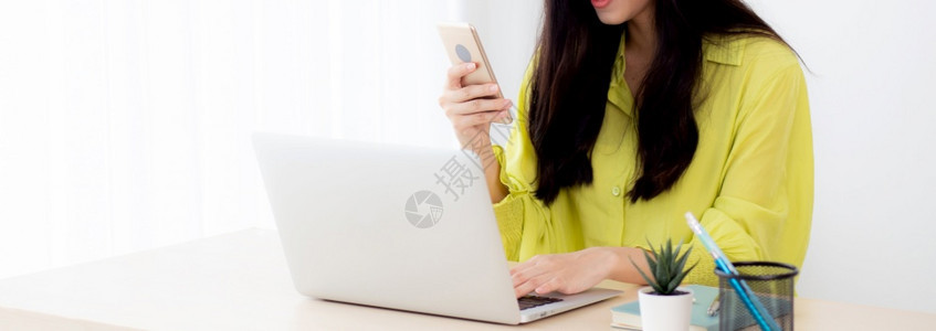在家办公室桌上在线使用笔记本电脑的年轻亚洲女商人在智能手机上寻找信息的自由女商人在桌子业和通信概念上打字的笔记本图片