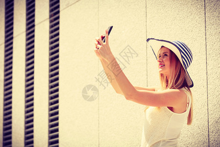 身戴太阳帽的年轻女用智能手机相拍摄自己用智能手机照相在阳光明媚的夏季日外拍摄在用智能手机拍自在外用智能手机拍自的年轻女图片