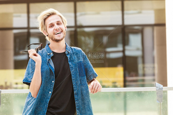 男子时装城市风格的服装概念穿着牛仔服男手提包在阳光明媚的一天握着太阳眼镜的嬉皮士在城市街头微笑的家伙图片