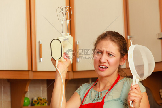 手拿着搅拌机的女主妇和头部筛子训练员围裙上的成熟女人在厨房玩得开心图片