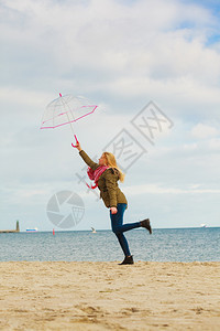 幸福享受寒冷的秋天气感受伟大的概念女人在海边滩上阳光明媚的白天和清蓝空上用透明的伞跳跃女人在海滩上用透明的伞跳跃图片