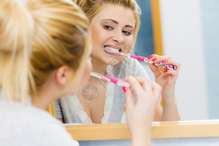 洗刷牙齿的女子洗刷牙金发女孩洗手间有牙刷的金发女孩看着镜子口腔卫生洗刷牙的女子在洗手间刷牙图片