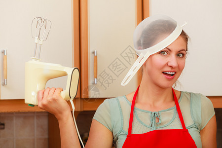 手持搅拌机的女主妇和头部筛子训练员围裙的年轻女人在厨房玩得开心的女主妇和搅拌机的女主妇在厨房玩得开心图片