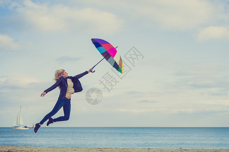 幸福享受寒冷的秋天气感受伟大的概念女人在海边滩上阳光明媚的白天和清蓝空上带着多彩的雨伞跳女人在海滩上跳图片