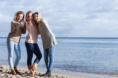 三个时髦的姐妹朋友在温暖的秋天气中穿着毛衣在阳光明媚的海滩上闲时间图片